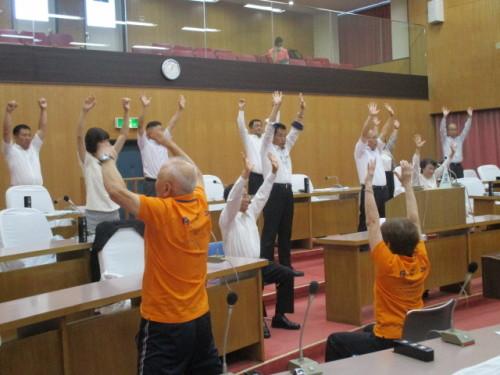 議会上の机の前で議員たちが両手を上に上げ体操をしている写真