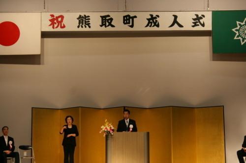 祝熊取町成人式と上に文字があり、奥には金屏風がある舞台で町長が話をしていて、横に立っている女性が手話の通訳をしている写真