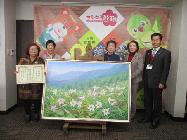 中央には大きな額縁に入った白い花の絵画が置かれていて、絵の周りに4人の女性、町長が並んでいる写真