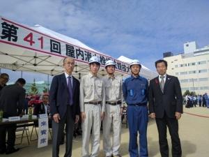 作業着を着て、ヘルメットをかぶった男性3名と、町長と、男性が横にならんでいる記念写真