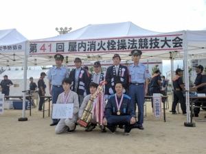 屋内消火栓操法競技大会で優勝したメンバーがトロフィーと賞状を持っている記念写真