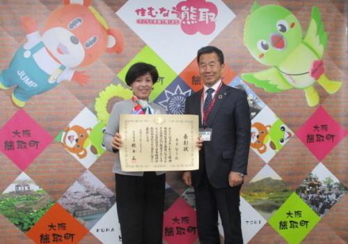 ジャンプ君とメジーナちゃんの描かれた壁の前で表彰状を持った本多智子氏と町長が並んでいる記念写真