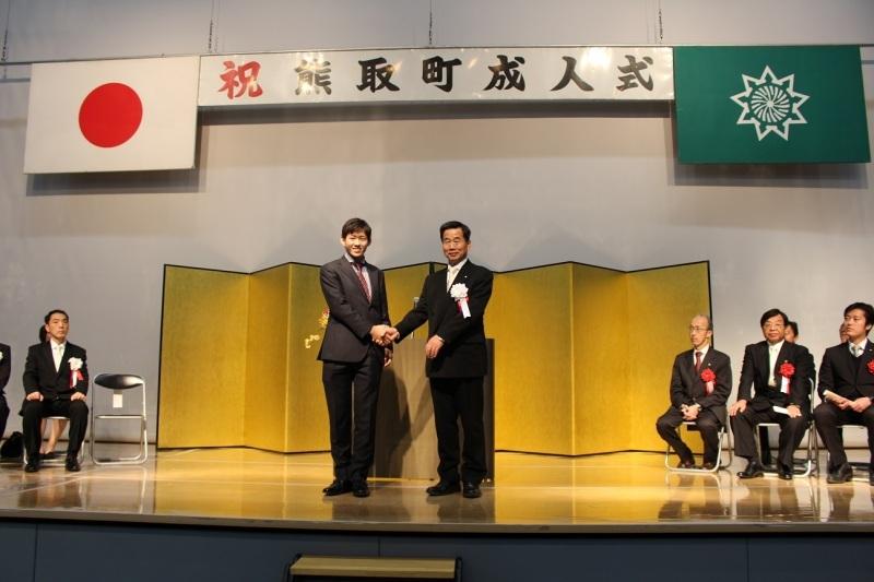 祝熊取町成人式と書かれた金屛風のある舞台上で町長と松井虎太郎さんが握手をしている写真