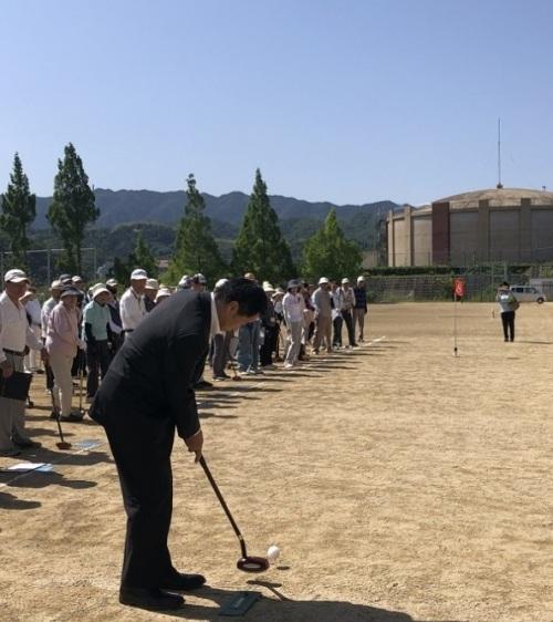 グラウンドでゴルフクラブを持ちボールを打つ町長とその様子を見る参加者の写真