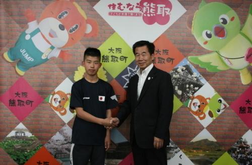 ジャンプ君とメジーナちゃんのが描かれた壁の前で渡邊康靖氏と町長が握手をして並んでいる写真