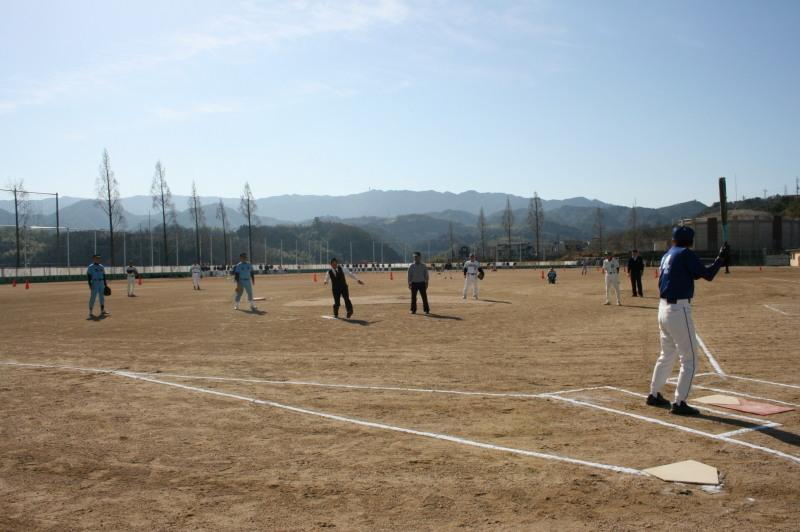 グラウンドの真ん中で白シャツにベストを着た町長がバッターに向かってボールを投げようとしている始球式の写真
