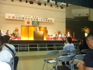 式典で、舞台の上に着席する関係者と、演台に立つ町長の写真