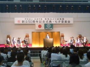 式典参加者が舞台上の演台に立つ町長を注目している写真