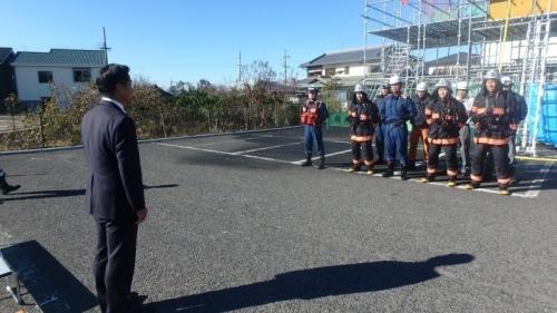 アスファルトの敷地に防火服を着た消防隊員が並んでいて、町長が隊員たちに向かって話をしている写真