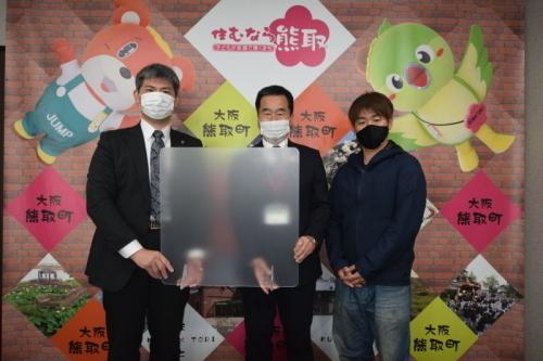 町長とスーツ姿の男性が二人でアクリル板を持って立ち、町長の横にパーカーを着た男性が並んで記念撮影している写真