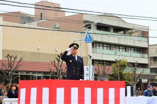 紅白の幕がある台の上で消防の制服姿の町長が右手を上にあげ敬礼をしている写真