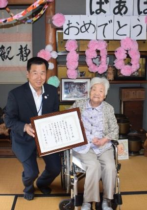 奥の壁に「ばあばおめでとう100」と文字が貼られていて、賞状を持った町長と小西スミヱさんが並んでいる記念写真