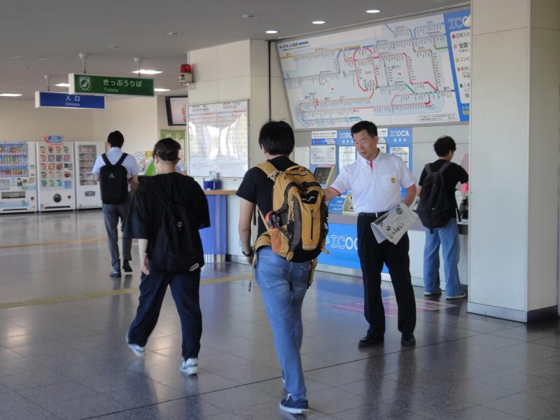 奥に切符販売機がある駅の構内で町長が通行人に広報紙を配布している写真