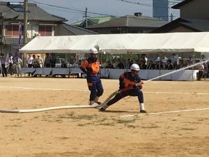 奥にはテントの下で座っている関係者たち、手前のグラウンドでは消防団員が長いホースから放水している様子が写っている写真