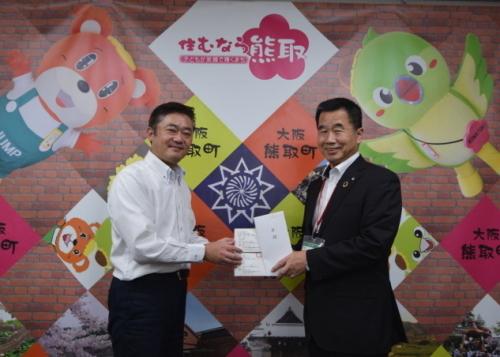 壁の前で佐古 員規代表取締役から町長が目録とマスク2箱を受け取っている写真