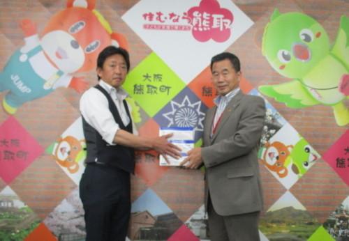 ジャンプ君とメジーナちゃんが描かれている壁の前で町長が西山 昌紀代表取締役からマスクを3箱受け取っている写真