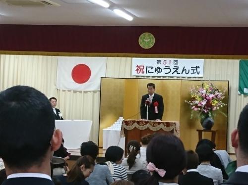祝入園式の文字と金屛風が置かれている舞台で来賓の花を胸につけた町長が祝辞を述べている写真