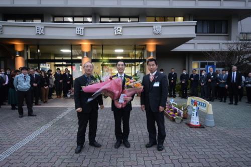 役場の入り口前に立っている沢山の職員の前で、花束を持つ中尾副町長と、町長と関係者の男性が並んでいる写真