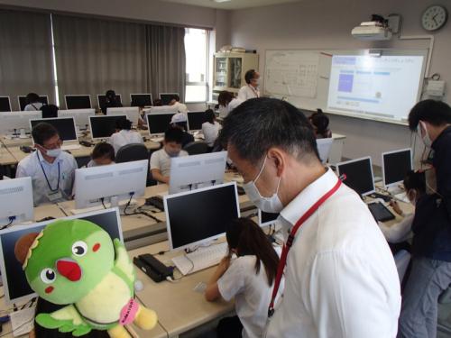 パソコンが設置されている教室内の手前にメジーナちゃんのぬいぐるみとマスク姿の町長、奥にはパソコンに向かいあっている生徒たちが写っている写真