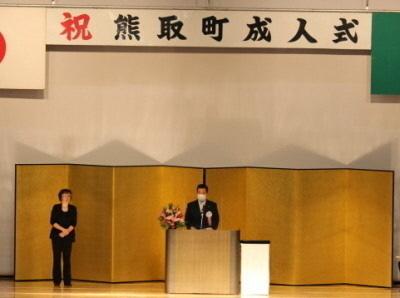 祝熊取町成人式と書かれた幕と金屛風が飾られた舞台の壇上で町長が話をしている写真