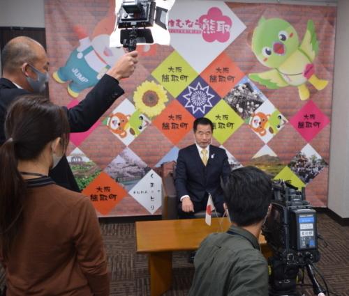 ライトで照らされ、カメラの前で椅子に座っている町長が話をしているJ:COM番組撮影の様子の写真