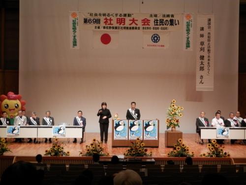 タスキをかけ演台の前で話す町長とその横で黒いスーツを着た女性が手話をしている写真