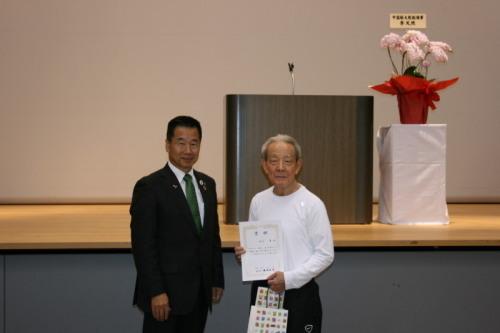 奥には胡蝶蘭の飾られた舞台があり、手前には賞状を持った男性、町長が並んで立っている写真