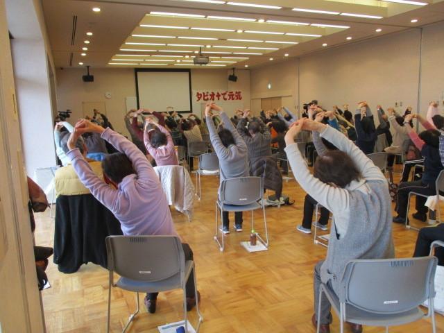 椅子に腰かけた参加者たちが両手を組み上に上げ、上半身を左に曲げている体操をしている様子を後ろから写した写真
