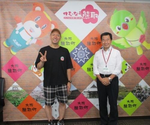 黒いTシャツにチェック柄の短パンを履いてピースサインをしている水本さんと白いYシャツ姿の町長が笑顔で並んで写っている写真