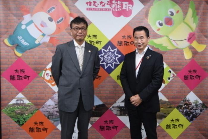 ジャンプ君とメジーナちゃんの絵が描かれている壁の前で和歌山大学学長と町長が並んでいる記念写真