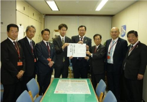 スーツを着た男性が8人並んでいて、中央の3人が白い紙に書かれた要望書を持っている写真