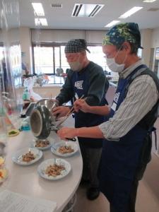 調理台の前に2人の男性が立っていて、鍋を裏返しながら皿に料理を取り分けている写真